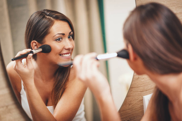 young smiling woman applying makeup in front of bathroom mirror - Foto, afbeelding