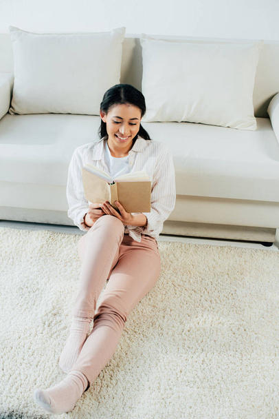 attractive latin woman seating reading book and smiling while sitting on floor at home - Φωτογραφία, εικόνα