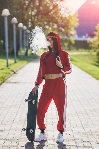 Vaping girl. Young woman with skateboard vape e-cig. Pretty young female in black hat, red clothing vape ecig, vaping device at the sunset. Toned image. Hip-hop style. - Photo, Image