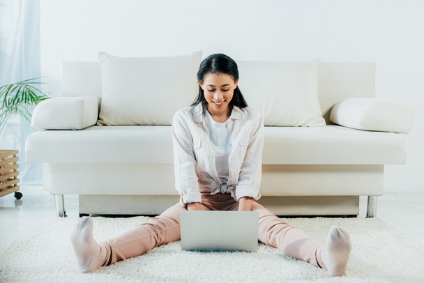 mujer latina alegre usando el ordenador portátil mientras está sentado en el suelo en casa
 - Foto, Imagen
