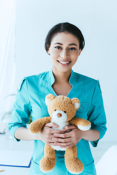 pretty latin doctor holding teddy bear while smiling at camera - Foto, Imagem