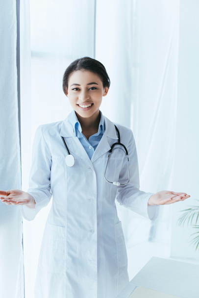 cheerful latin doctor with stethoscope showing welcome gesture while smiling at camera - Foto, Imagen