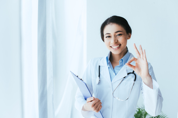 schöne lateinische Ärztin lächelt in die Kamera, während sie okay Geste zeigt und Klemmbrett hält - Foto, Bild