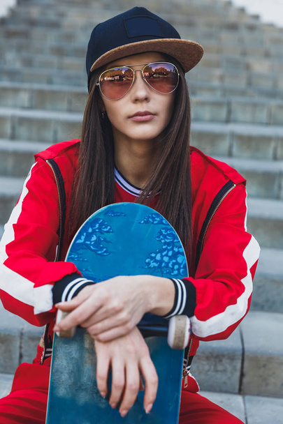 Portrait of young female wearing black hat, red clothing skateboarder holding her skateboard. Woman with skating board looking at camera outdoors. - 写真・画像