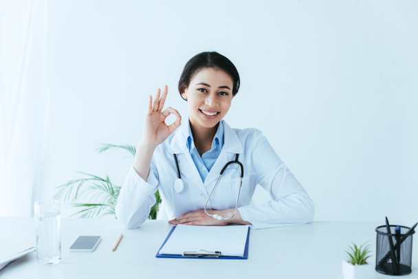 schöne lateinische Frau zeigt okay Geste und lächelt in die Kamera, während sie am Arbeitsplatz im Krankenhaus sitzt - Foto, Bild