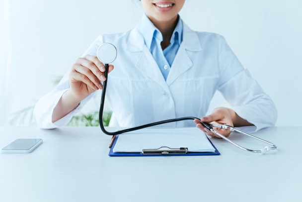 inyección recortada de joven médico latino sosteniendo estetoscopio mientras está sentado en el lugar de trabajo
 - Foto, imagen