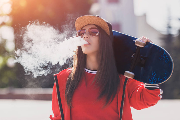 V, holka. Mladá žena se skateboard s e-cigem. Hezká mladá žena v černém klobouku, červený oděv, vpikadlo, zařízení při západu slunce. Tónový obraz. Hip-hop styl. - Fotografie, Obrázek
