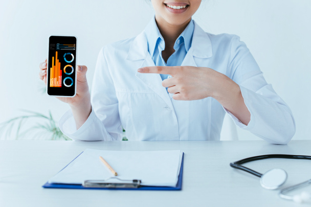 partial view of smiling latin doctor pointing with finger at smartphone with infographics on screen - Photo, Image