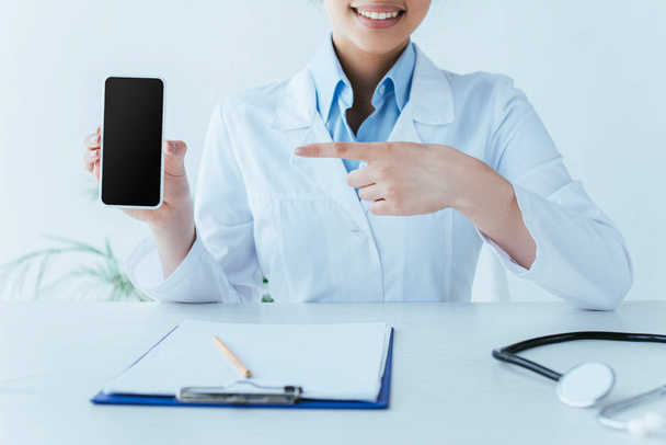 partial view of smiling latin doctor pointing with finger at smartphone with blank screen - Photo, Image