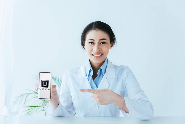 KYIV, UKRAINE - APRIL 26, 2019: Beautiful latin doctor pointing with finger at smartphone with  Uber app on screen. - 写真・画像