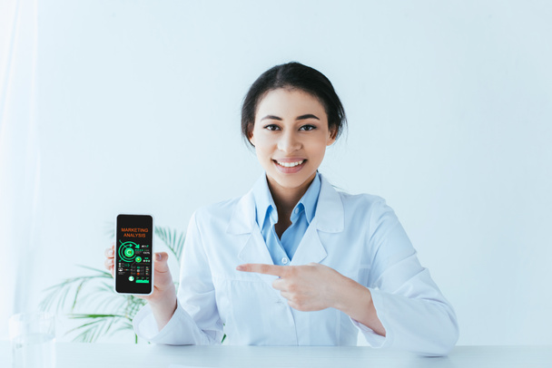 молодий латинський лікар вказує пальцем на смартфон з маркетинговим аналізом на екрані
 - Фото, зображення