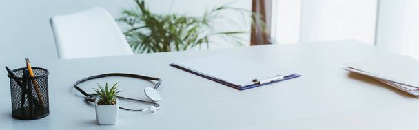 panoramic shot of clipboard with blank paper, stethoscope and green potted plant on desk in hospital - Photo, Image
