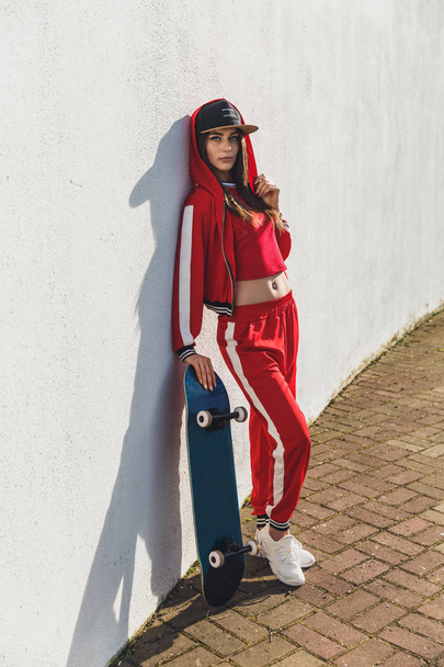 Portrait of young female wearing black hat, red clothing skateboarder holding her skateboard. Woman with skating board looking at camera outdoors. - Photo, Image