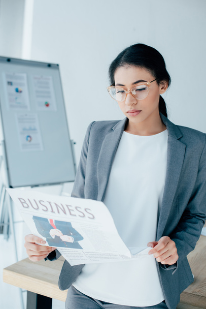 attentive latin businesswoman in glasses reading business newspaper in office - Photo, Image