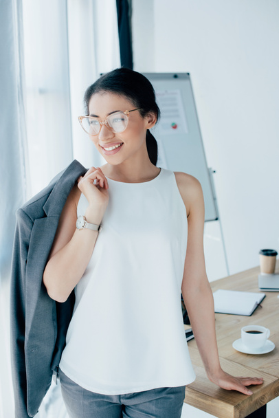 pretty latin businesswoman standing near workplace and looking away in office - Photo, Image
