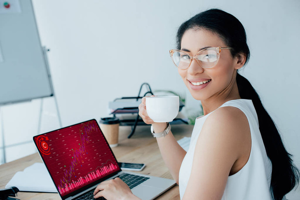 sorridente latino empresária usando laptop com infográficos na tela enquanto segurando xícara de café e sorrindo para a câmera
 - Foto, Imagem