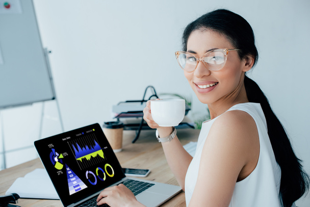 χαμογελαστή Λατινική επιχειρηματίας κρατώντας κούπα καφέ, ενώ χρησιμοποιεί φορητό υπολογιστή με γραφήματα και διαγράμματα στην οθόνη - Φωτογραφία, εικόνα