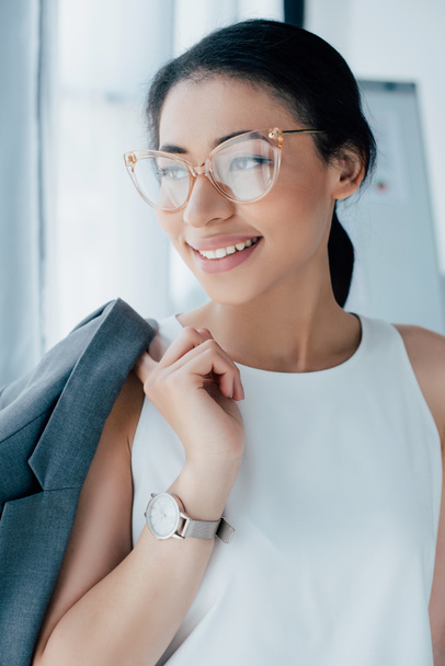 beautiful latin businesswoman in glasses smiling and looking away in office - Foto, Bild