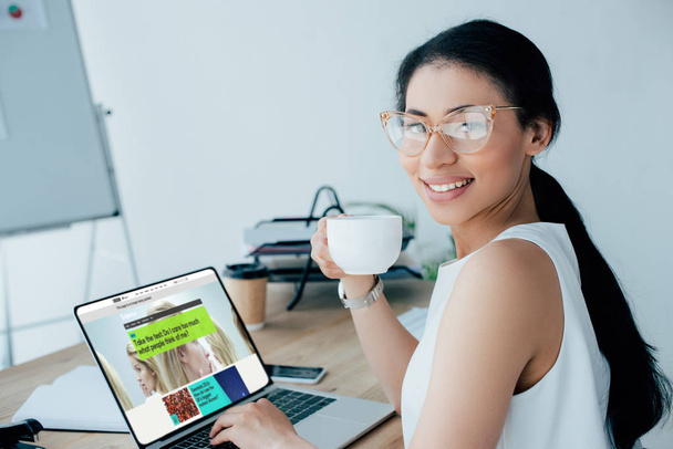 KYIV, UKRAINE - APRIL 26, 2019: Attractive latin businesswoman holding coffee cup while using laptop with BBC Science website on screen.  - 写真・画像