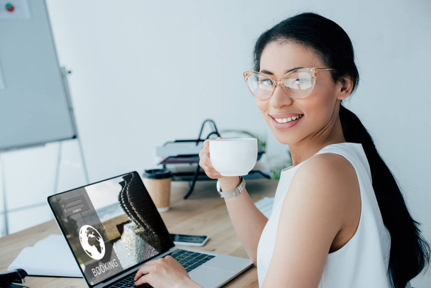 sorridente latino empresária segurando xícara de café ao usar laptop com site de reserva na tela
 - Foto, Imagem