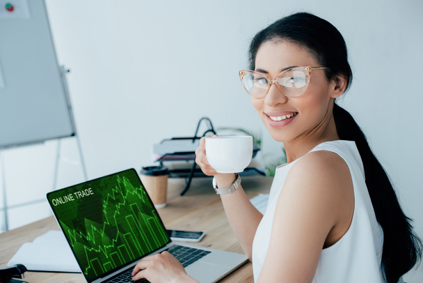 smiling latin businesswoman using laptop with online trade website on screen while holding coffee cup - Фото, изображение