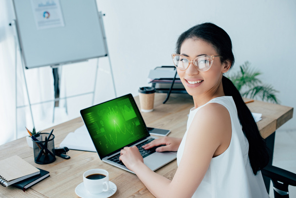 attrayant latine femme d'affaires dans des lunettes à l'aide d'un ordinateur portable avec des graphiques et des graphiques à l'écran tout en souriant à la caméra
 - Photo, image