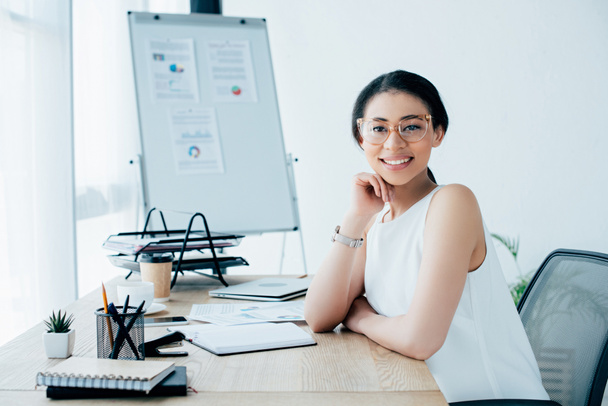 cheerful latin businesswoman in glasses smiling at camera while sitting at workplace in office - Foto, Imagen