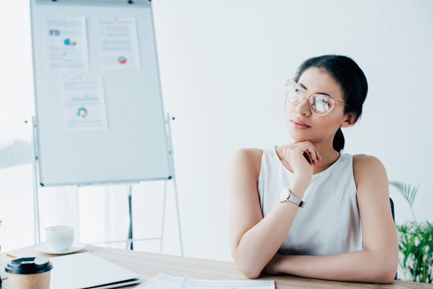 mujer de negocios latina soñadora con gafas mirando hacia otro lado mientras está sentada en el lugar de trabajo
 - Foto, imagen