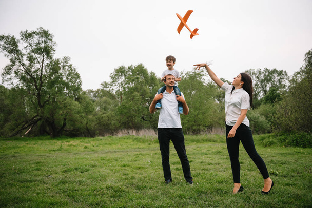 Ojciec, matka i syn bawią się zabawkowym samolotem w parku. przyjazna rodzina. Ludzie bawią się na świeżym powietrzu. Obraz wykonany na tle parku i błękitnego nieba. koncepcja szczęśliwej rodziny. - Zdjęcie, obraz