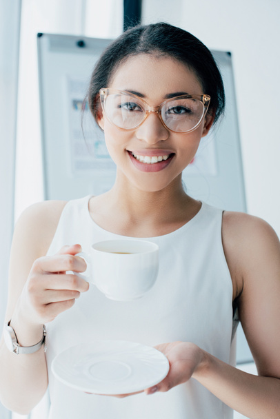 cheerful businesswoman in glasses holding coffee cup and smiling at camera - Foto, afbeelding