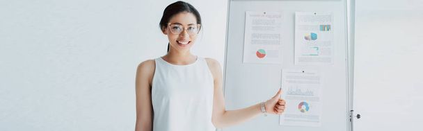 panoramic shot of cheerful latin businesswoman showing thumb up while standing near flipchart with infographics - Фото, зображення