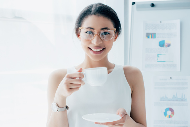 hermosa mujer de negocios latina en vasos sosteniendo taza de café mientras sonríe a la cámara
 - Foto, imagen