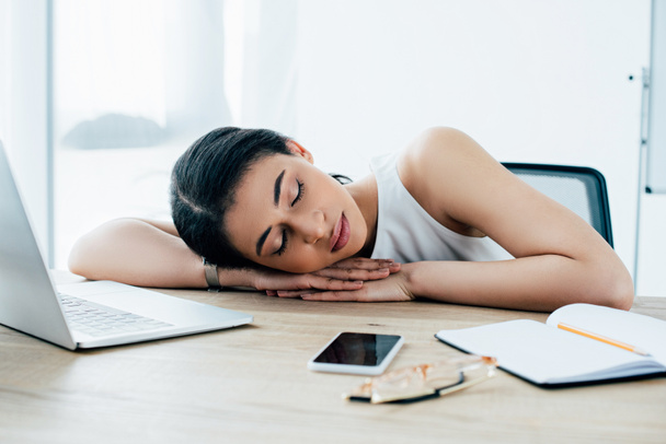 esausta donna d'affari latina che dorme sul posto di lavoro vicino al notebook e allo smartphone con schermo bianco
 - Foto, immagini
