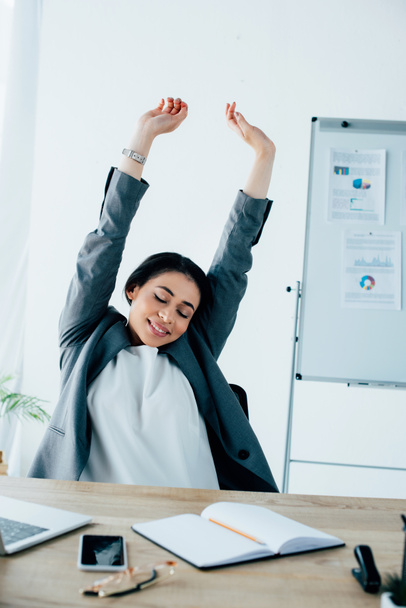 exhausted latin businesswoman stretching with closed eyes while sitting at workplace - Foto, immagini