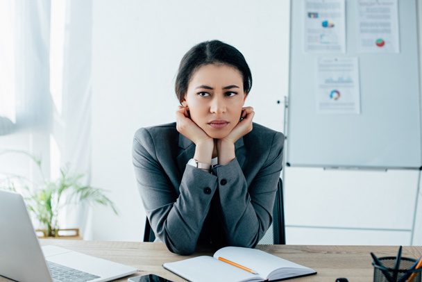 upset latin businesswoman looking away while sitting at workplace in office - 写真・画像