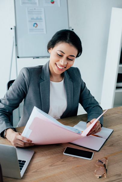 mujer de negocios latina sonriente sosteniendo una carpeta de papel mientras está sentada en el lugar de trabajo cerca de un teléfono inteligente con pantalla en blanco
 - Foto, Imagen