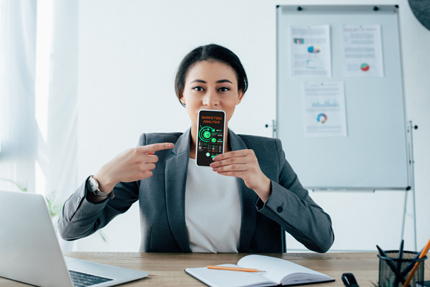όμορφη Λατινική επιχειρηματίας που δείχνει το smartphone με την εφαρμογή ανάλυσης μάρκετινγκ στην οθόνη - Φωτογραφία, εικόνα