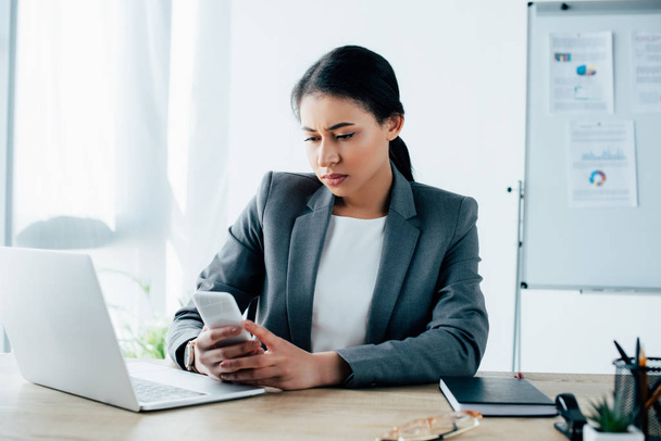 serious latin businesswoman using smartphone while sitting at workplace near laptop - Foto, imagen