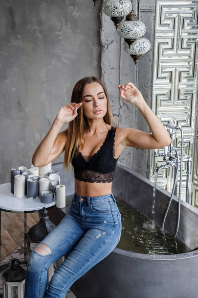 Closeup portrait of beauty young woman in black lingerie and blue jeans posing at modern bathroom interior - Photo, Image