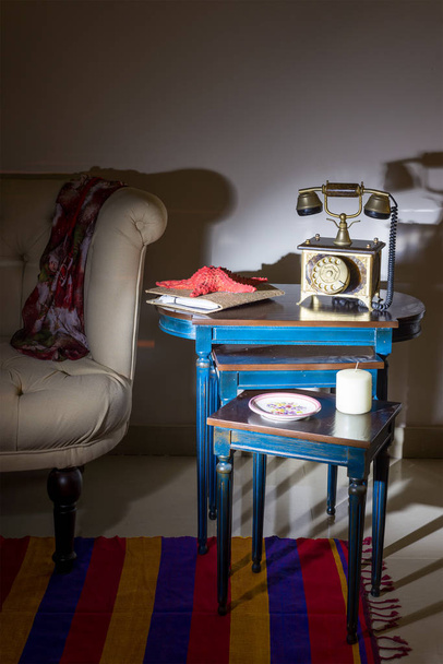 Innenaufnahme eines alten goldenen Telefons, weißer Kerze, rotem Seestern und bunt verziertem Teller über drei verschachtelten Tischen mit blauen Beinen über einer weißen Wand und cremefarbenem Sessel mit grellem Licht - Foto, Bild