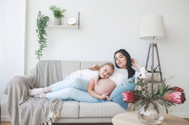 妊娠中の女性と彼女の小さな娘は、屋内で楽しみを持っています。マタニティ。小さなかわいい娘との赤ちゃんの出産を待っている若い母親。家族一緒に. - 写真・画像