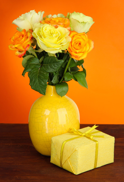Beautiful bouquet of roses in vase with gift on table on orange background - Foto, immagini