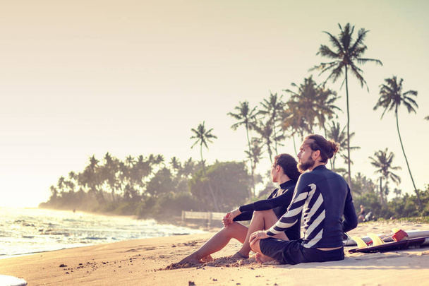 Jovens e belos amigos amorosos estão sentados na areia junto ao oceano com pranchas de surf
 - Foto, Imagem