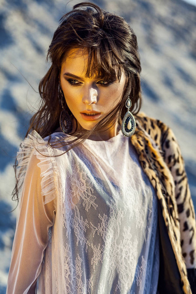 attractive brunette woman in black body and translucent beach cover up with fur coat posing on sandy beach at sunset  - Fotoğraf, Görsel