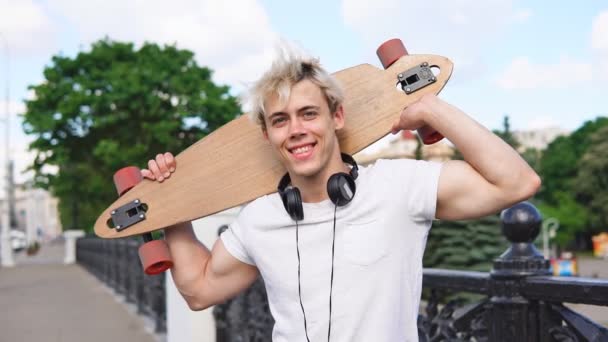 Vrolijke blonde man met hoofdtelefoon die Longboard achter het hoofd houdt en glimlacht - Video
