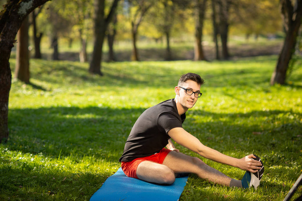 Jongeman met een bril die buiten yoga traint. Sportieve man maakt ontspannen oefening op een blauwe yoga mat, in het park. Kopieerruimte - Foto, afbeelding