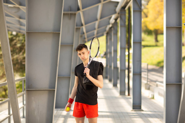 The man poses outdoors with a tennis racket and a ball. A towel is hanging on his shoulder. Sport concept - Foto, Bild