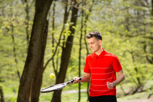 Mies punaisessa t-paidassa poseeraa tennismailan ja pallon kanssa vihreän puiston taustalla. Urheilukäsite
 - Valokuva, kuva