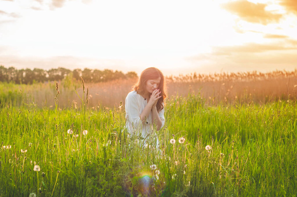 Το κορίτσι έκλεισε τα μάτια της, προσευχόταν σε ένα χωράφι κατά τη διάρκεια του όμορφου ηλιοβασιλέματος. Τα χέρια διπλωμένα στο σκεπτικό της προσευχής για την πίστη - Φωτογραφία, εικόνα