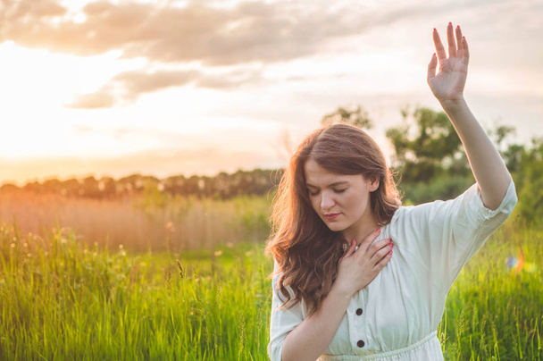 Menina fechou os olhos, rezando em um campo durante o belo pôr do sol. Mãos dobradas no conceito de oração pela fé
 - Foto, Imagem